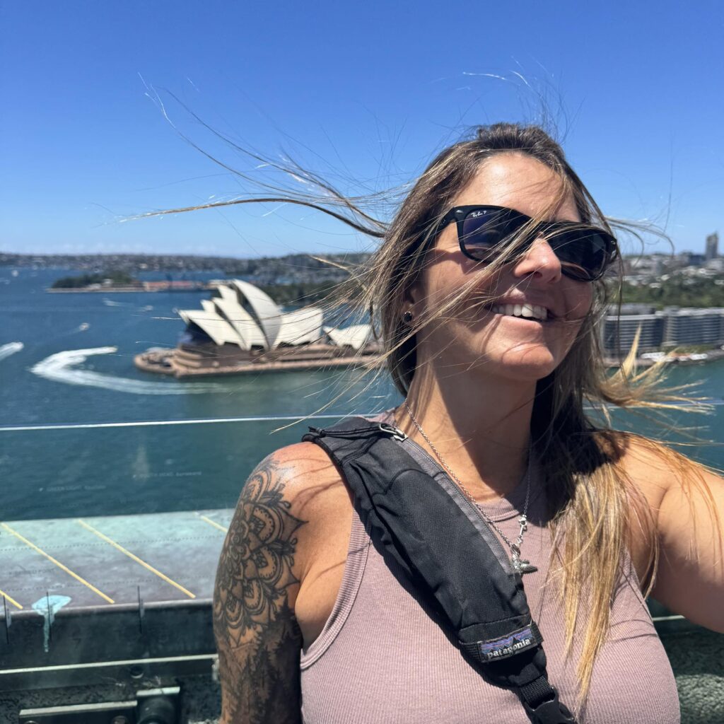 Woman with long hair smiles against a backdrop of Sydney's vibrant harbor, featuring the iconic Sydney Opera House and blue water.