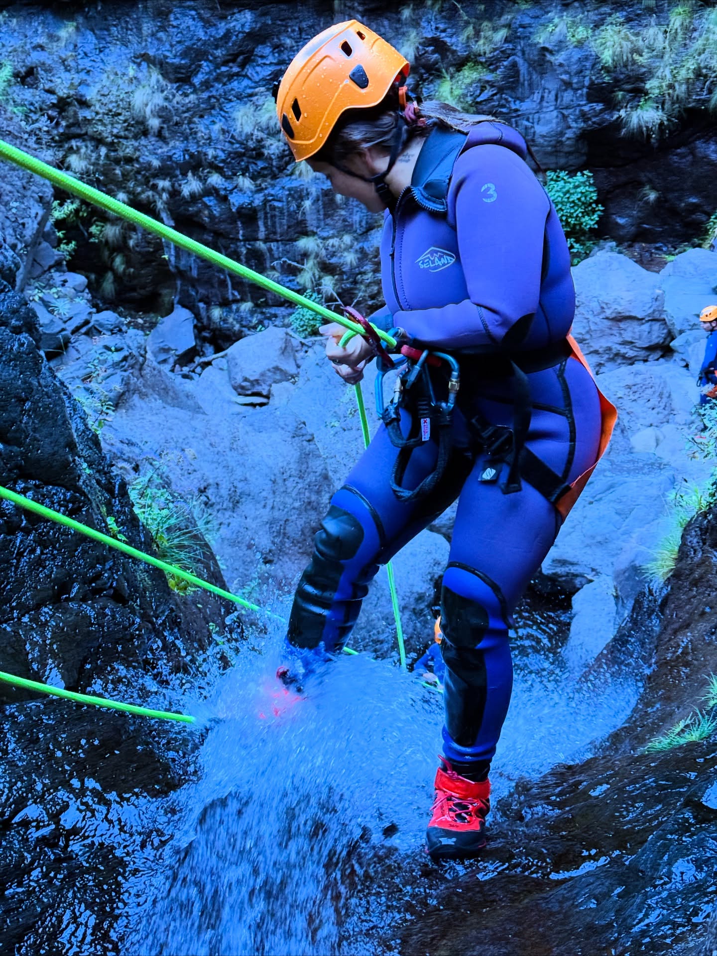A woman in a wetsuit and helmet rappels down a rocky canyon, water splashing around her feet. Green ropes and a rocky backdrop highlight the outdoor adventure.