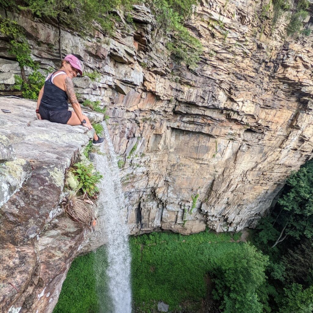 A person sits on the edge of a rocky cliff, overlooking a waterfall cascading into lush green vegetation below. The scene captures the beauty of nature and adventure.