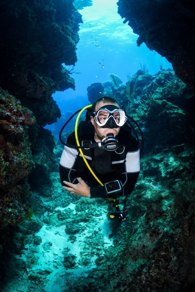 Diver exploring a coral reef through underwater rock formations, showcasing marine life and vibrant ocean colors. Relevant for marine biology or diving topics.