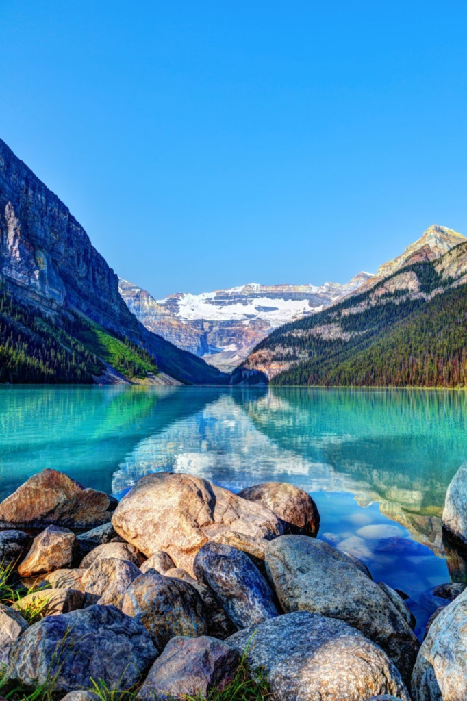 Clear turquoise lake reflects majestic mountains and clear blue sky, framed by rocky shores and lush forests. Ideal for nature enthusiasts.