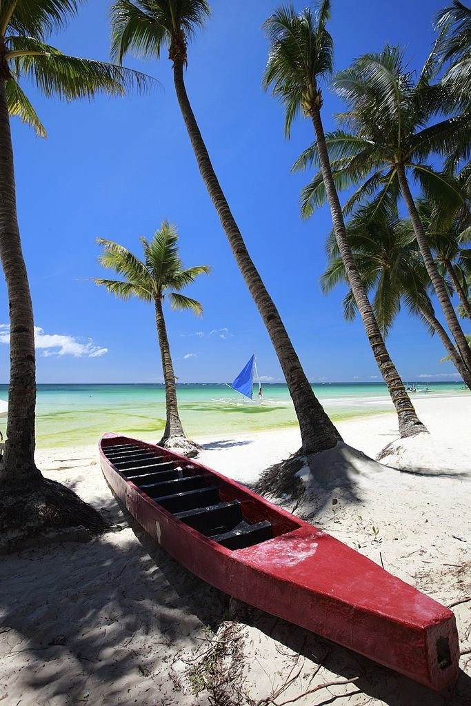 A red canoe rests on sandy beach under palm trees, with a blue sailboat in the background against a clear sky and tranquil turquoise waters.