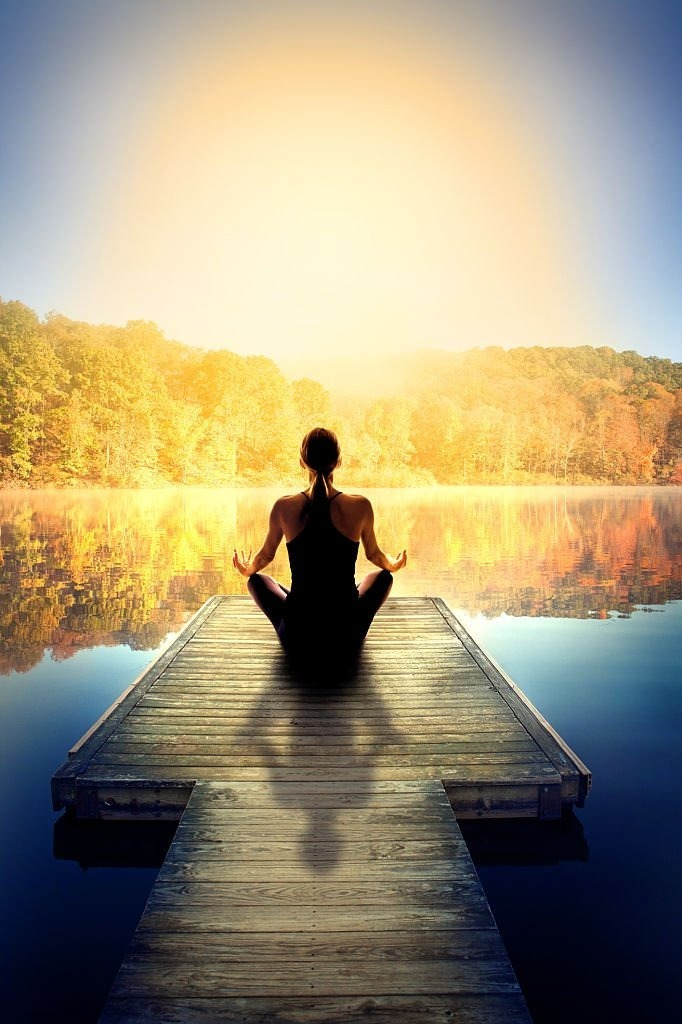 Person meditating on a wooden dock by a serene lake, surrounded by autumn foliage, under a softly glowing sky.