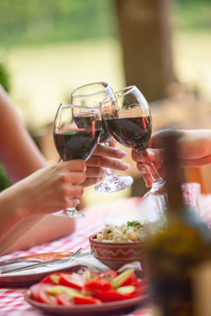 Friends toasting with glasses of red wine at a table laid with food, celebrating together in a warm, outdoor setting.